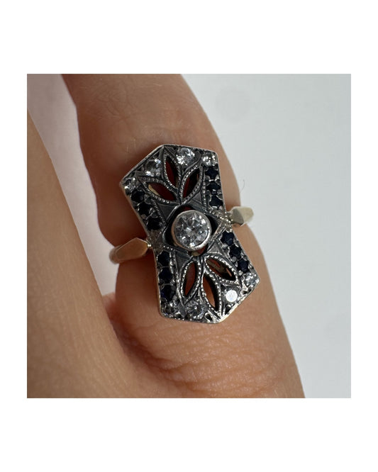 טבעת זהב 14 קראט בעיצוב אר-דקו גיאומטרי, משובצת יהלום מרכזי ומסגרת של אבני ספיר כהות ויהלומים נוצצים.