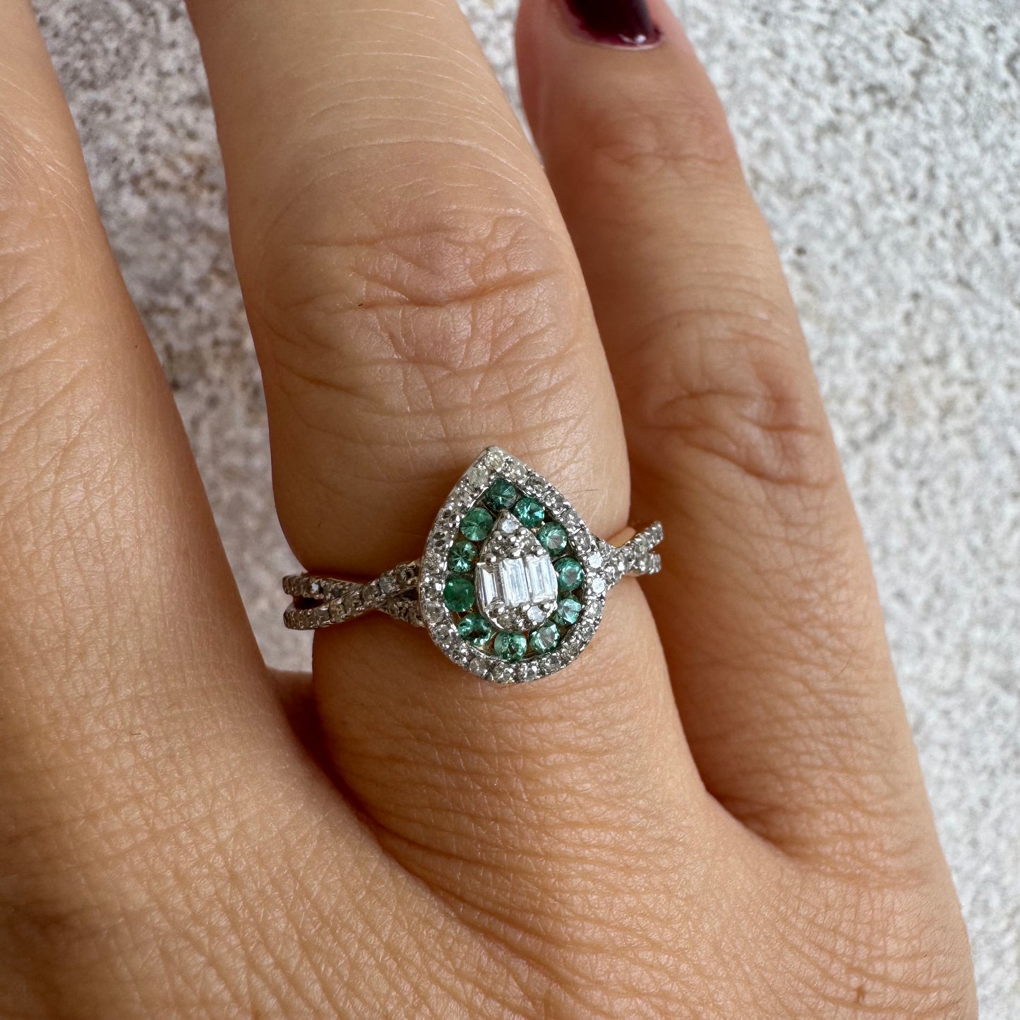 טבעת זהב צהוב 14 קראט בעיצוב טיפה (pear shape), משובצת יהלומים לבנים בשיבוץ הילה כפולה (double halo) ואבני אמרלד קטנות המקיפות מרכז ייחודי של יהלומים בחיתוך באגט