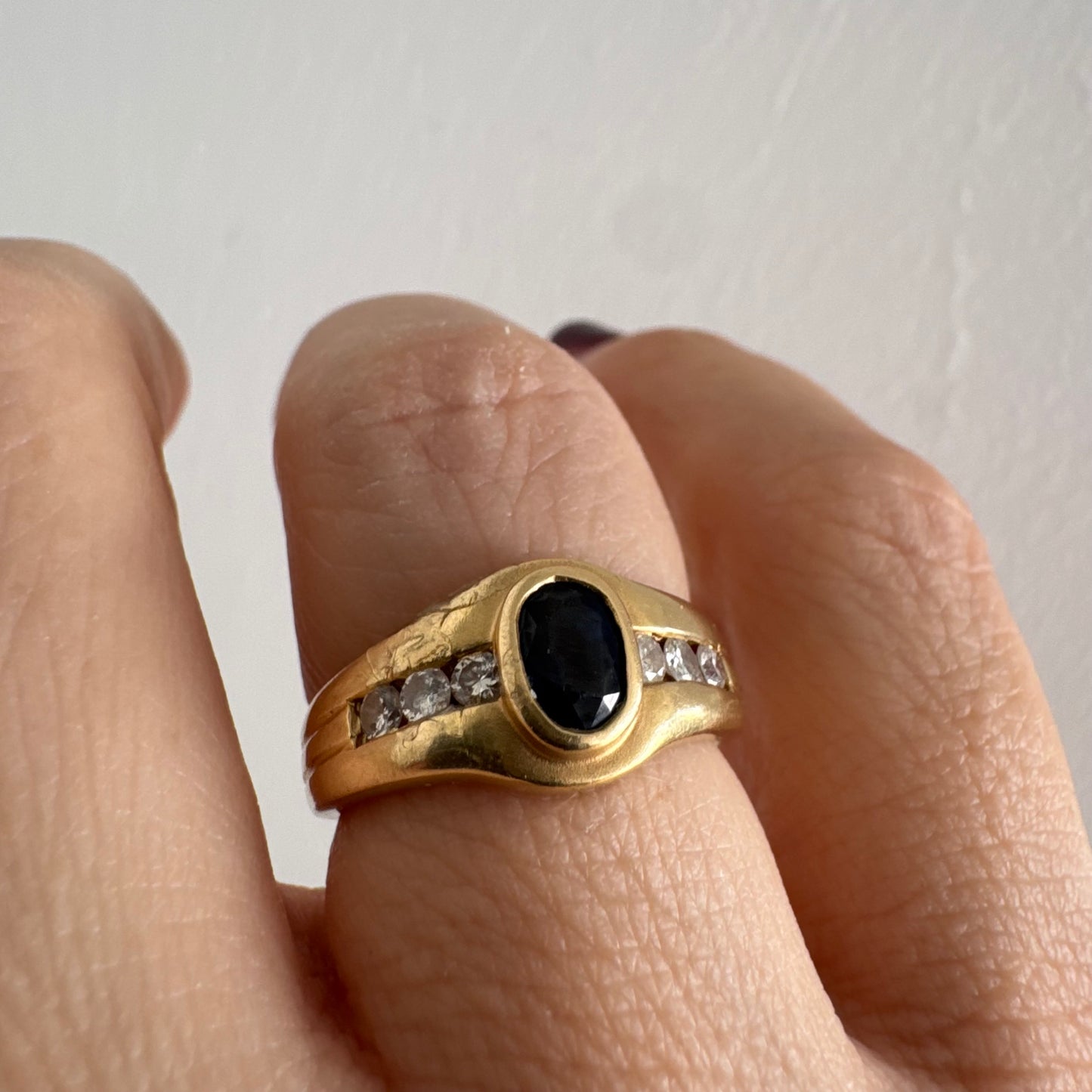 טבעת זהב צהוב 18 קראט משובצת אבן ספיר אובלית בגוון כחול עמוק, במסגרת חלקה ומודגשת, ומשני צדדיה שורת יהלומים משובצים בתעלה.