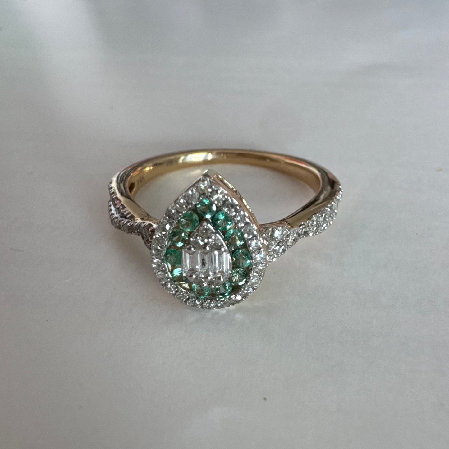 טבעת זהב צהוב 14 קראט בעיצוב טיפה (pear shape), משובצת יהלומים לבנים בשיבוץ הילה כפולה (double halo) ואבני אמרלד קטנות המקיפות מרכז ייחודי של יהלומים בחיתוך באגט