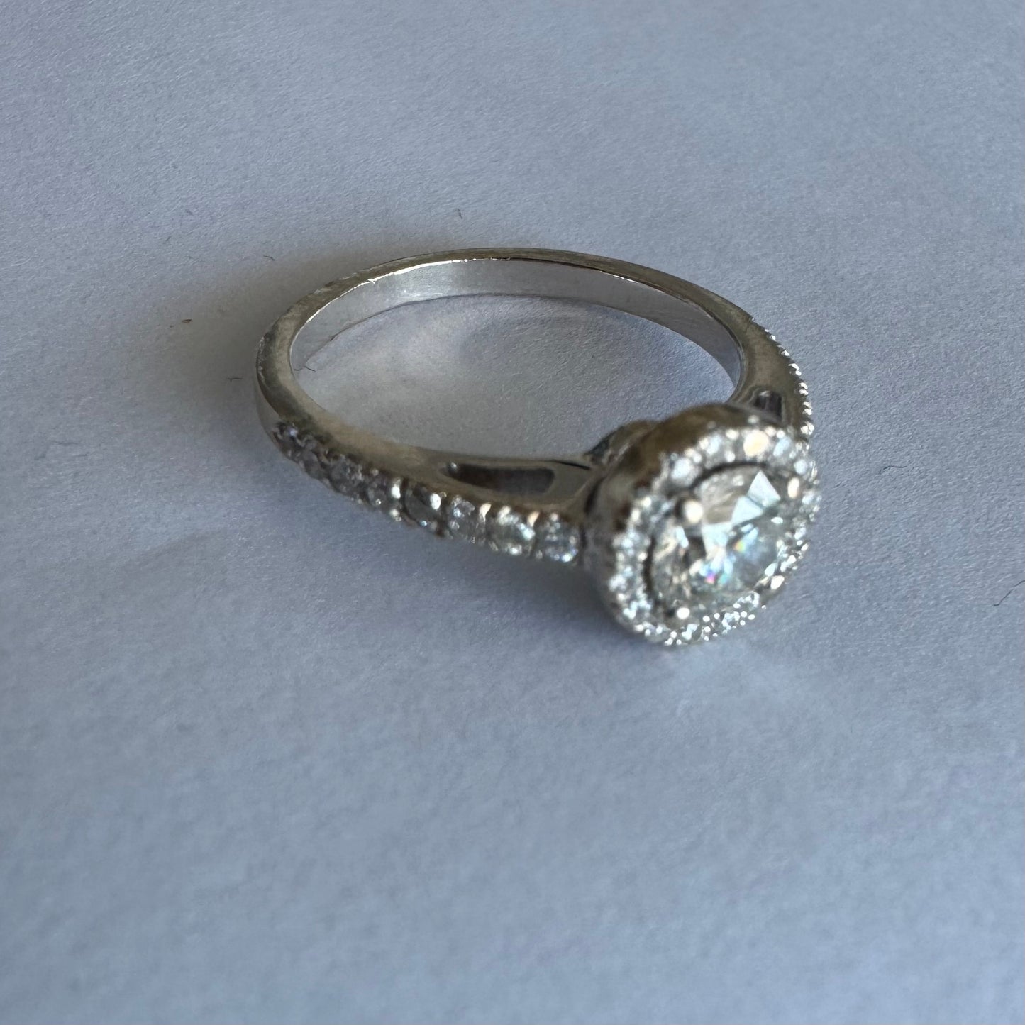 טבעת זהב לבן 14 קראט משובצת יהלום מרכזי עגול והילה נוצצת של יהלומים קטנים, עם יהלומים נוספים על החישוק.