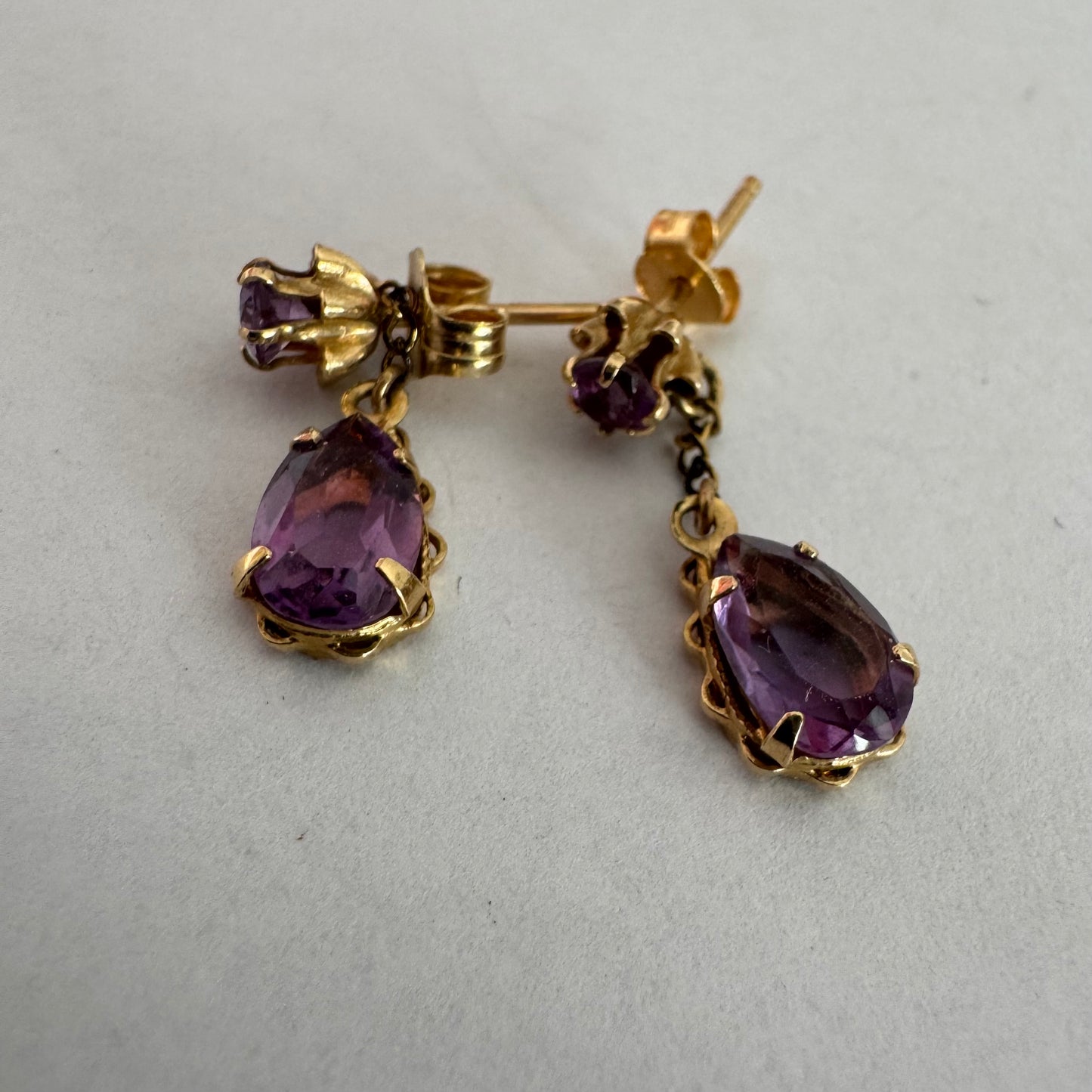 עגילים זהב צהוב 14 קראט משובצים אבני אמטיסט בגוון סגול עמוק בחיתוך טיפה, בשילוב אבן עליונה תואמת ועיטור שרשרת עדינה המחברת ביניהן.
