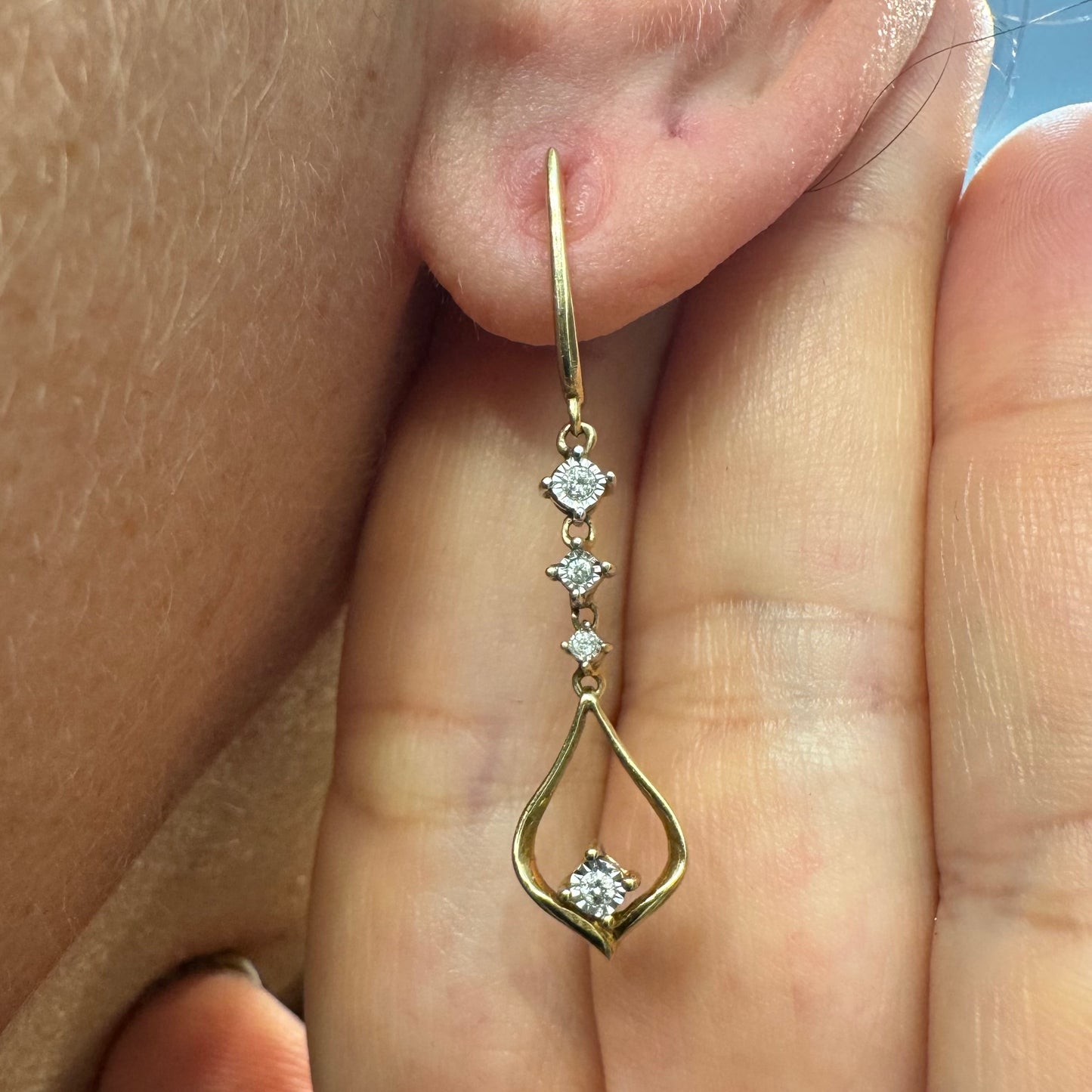 עגילי זהב 14 קראט תלויים בעיצוב עדין ומודרני, משובצים יהלומים לבנים לאורך החלק המרכזי ובטיפה פתוחה בתחתית משובץ יהלום נוסף.