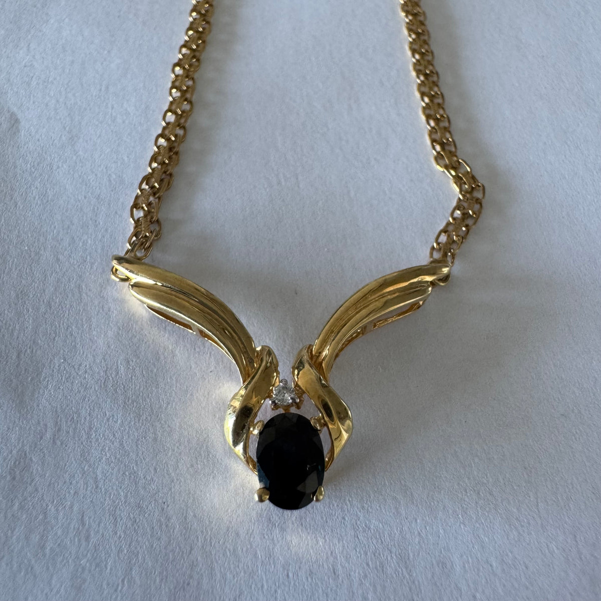 14K gold necklace featuring a flowing pendant design, set with a dark blue center Sapphire and a small Diamond above.