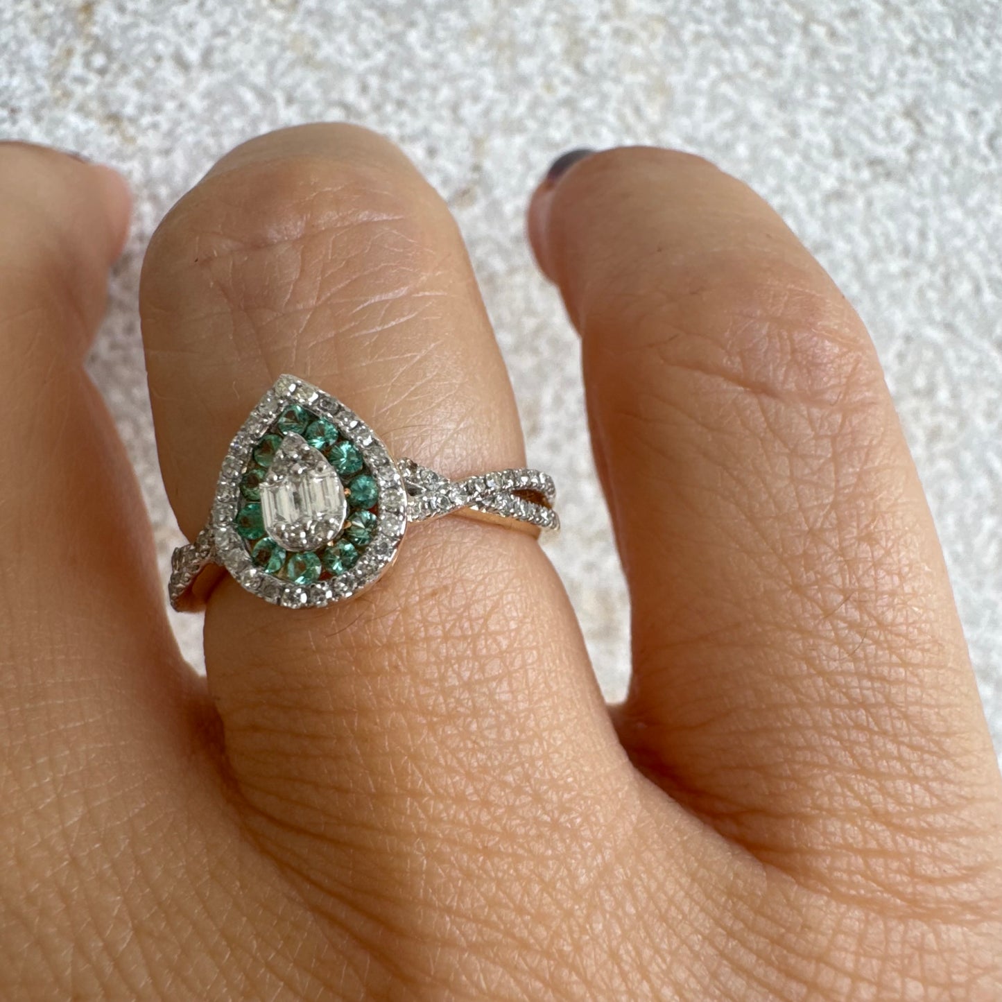 טבעת זהב צהוב 14 קראט בעיצוב טיפה (pear shape), משובצת יהלומים לבנים בשיבוץ הילה כפולה (double halo) ואבני אמרלד קטנות המקיפות מרכז ייחודי של יהלומים בחיתוך באגט