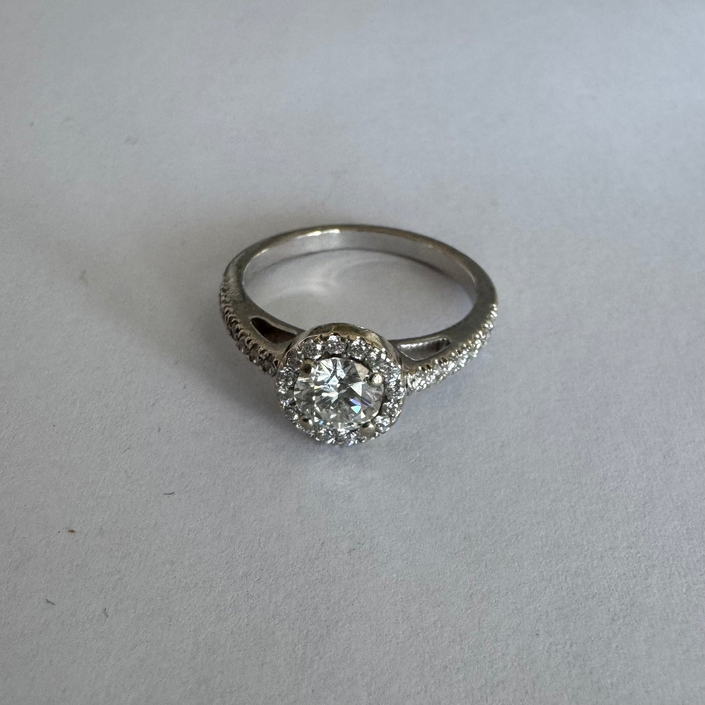 טבעת זהב לבן 14 קראט משובצת יהלום מרכזי עגול והילה נוצצת של יהלומים קטנים, עם יהלומים נוספים על החישוק.