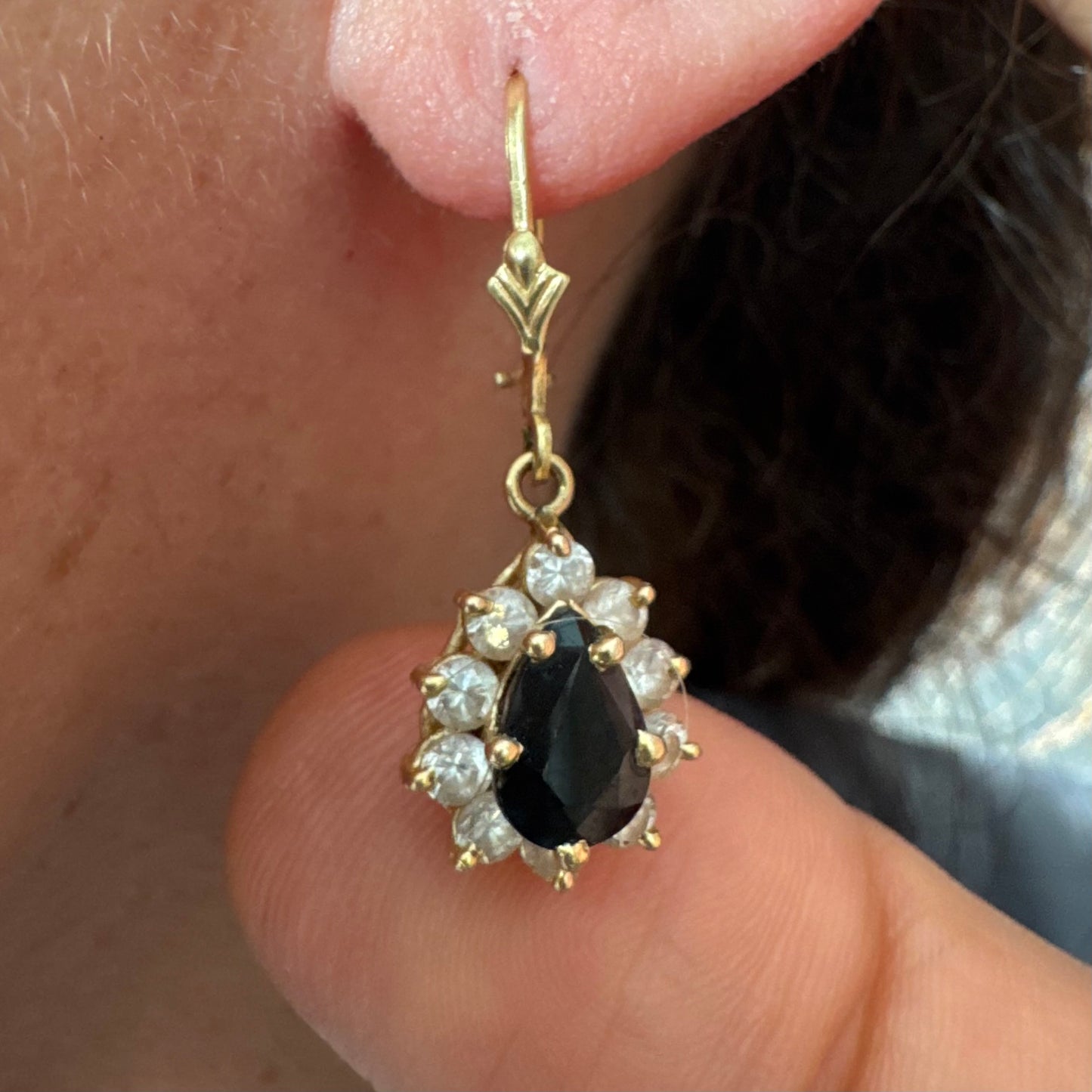 עגילי זהב צהוב 14 קראט בעיצוב קלאסי ומרשים, משובצים אבן ספיר בגוון כחול כהה בחיתוך טיפה ומסביבה אבנים שקופות עם הילה נוצצת שמוסיפה אור וזוהר לכל תנועה.