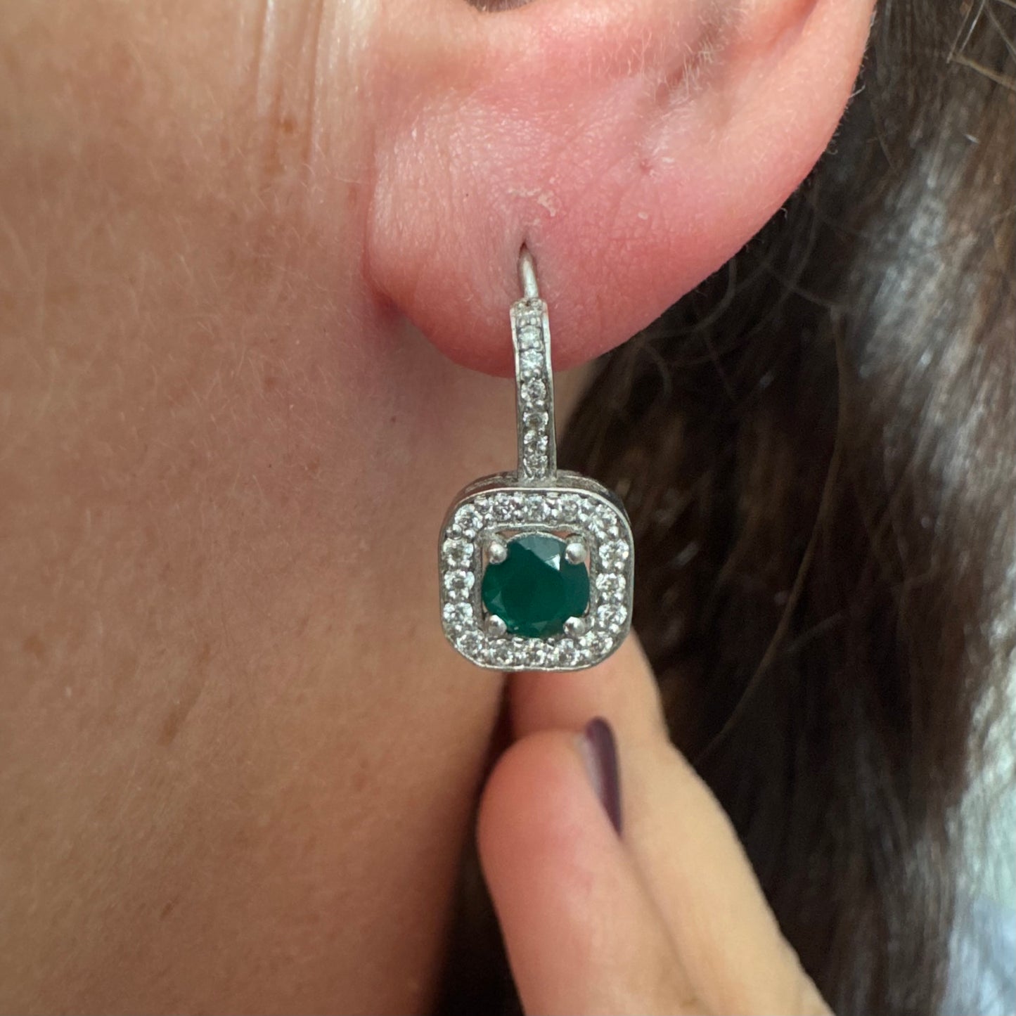 עגילי זהב לבן 14 קראט בעיצוב אלגנטי ונקי, משובצים אבן אמרלד מרכזית במראה עשיר ועמוק, מוקפת הילה מנצנצת של יהלומים.