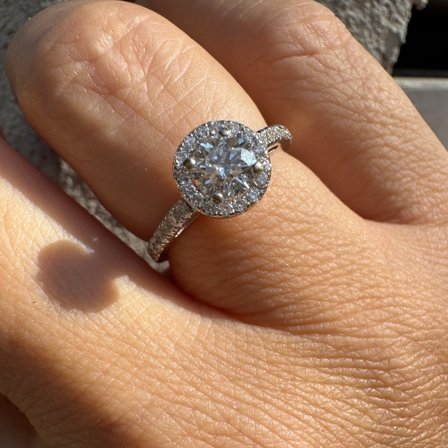 טבעת זהב לבן 14 קראט משובצת יהלום מרכזי עגול והילה נוצצת של יהלומים קטנים, עם יהלומים נוספים על החישוק.