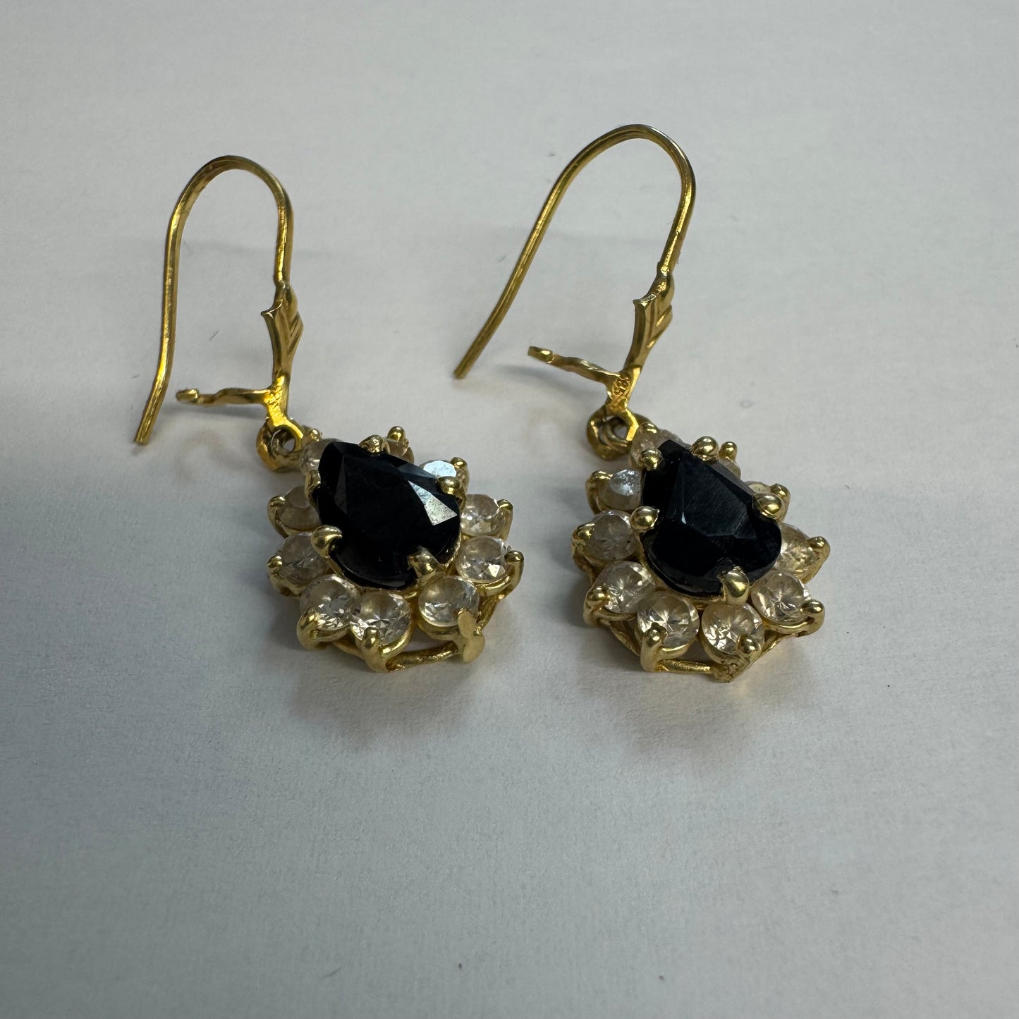 עגילי זהב צהוב 14 קראט בעיצוב קלאסי ומרשים, משובצים אבן ספיר בגוון כחול כהה בחיתוך טיפה ומסביבה אבנים שקופות עם הילה נוצצת שמוסיפה אור וזוהר לכל תנועה.