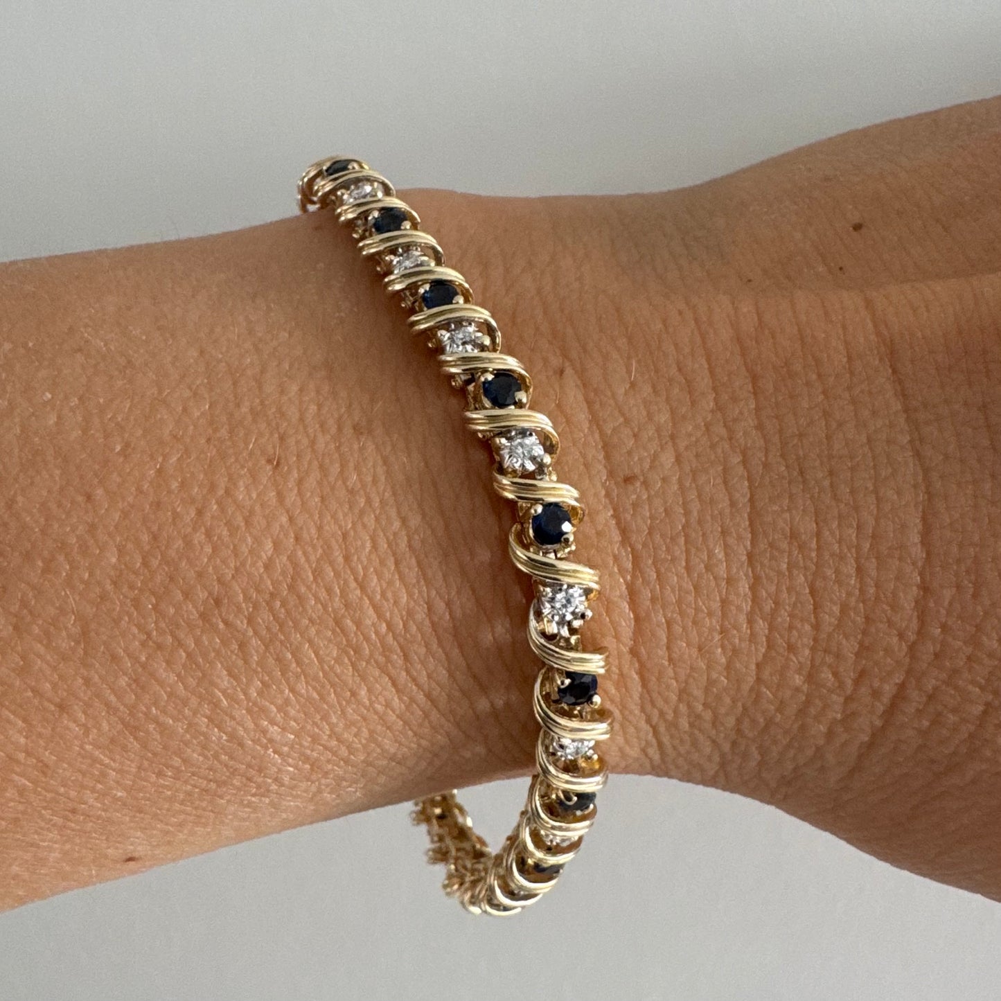 צמיד זהב צהוב 14 קראט משובץ אבני ספיר כחולות ויהלומים, המשובצים בעיצוב זורם עם קווים מעוגלים העוטפים כל אבן.