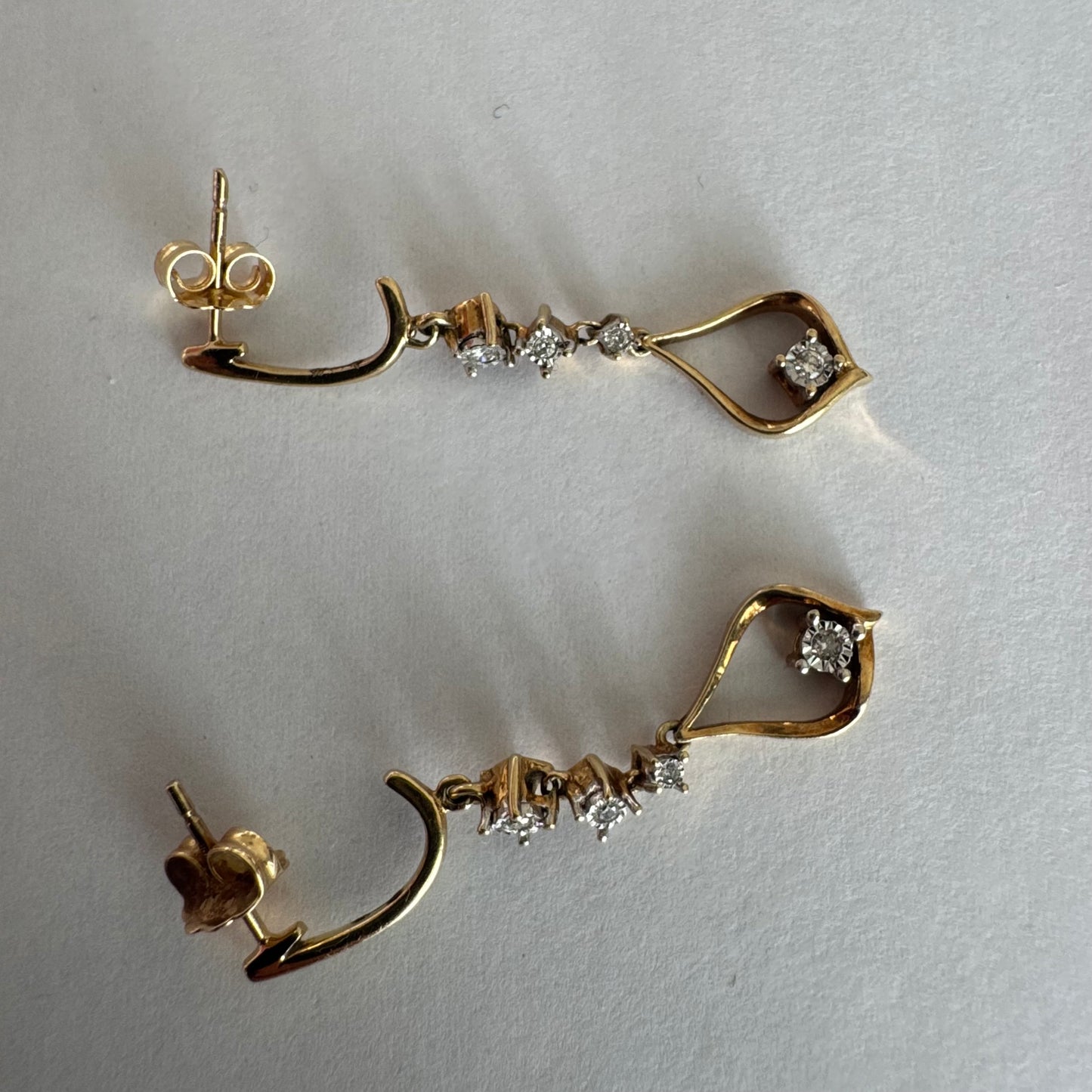 עגילי זהב 14 קראט תלויים בעיצוב עדין ומודרני, משובצים יהלומים לבנים לאורך החלק המרכזי ובטיפה פתוחה בתחתית משובץ יהלום נוסף.