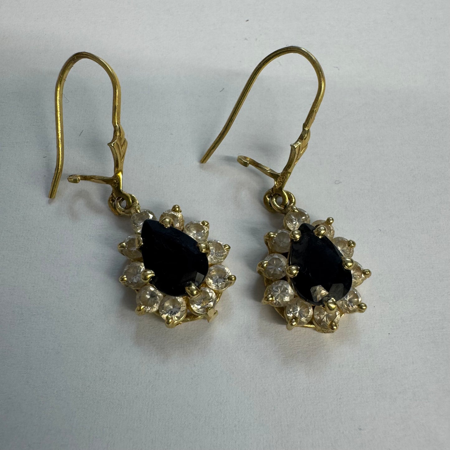 עגילי זהב צהוב 14 קראט בעיצוב קלאסי ומרשים, משובצים אבן ספיר בגוון כחול כהה בחיתוך טיפה ומסביבה אבנים שקופות עם הילה נוצצת שמוסיפה אור וזוהר לכל תנועה.