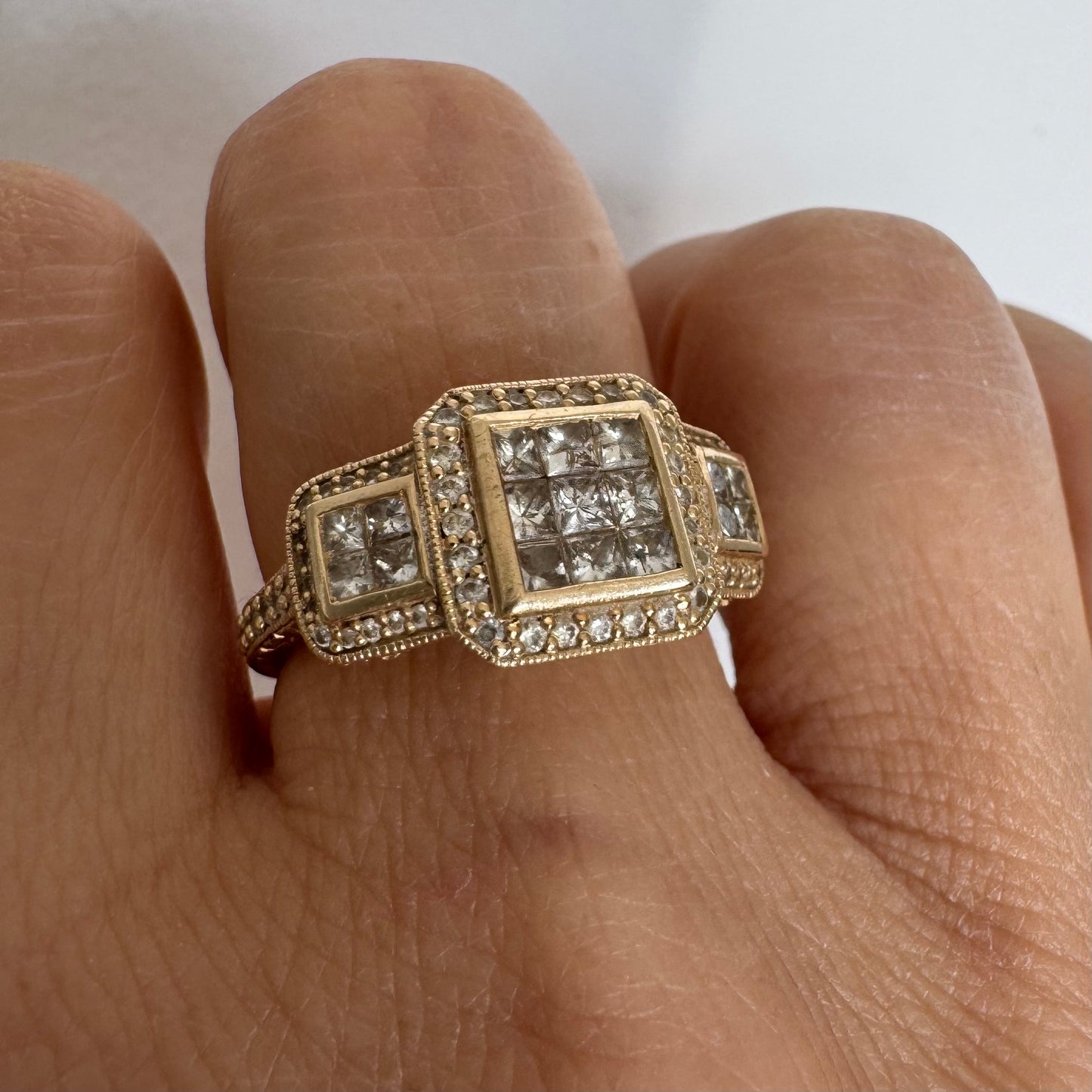 טבעת זהב צהוב 14 קראט משובצת יהלומים בחיתוך פרינסס במרכז, מוקפת הילה של יהלומים צדדיים.