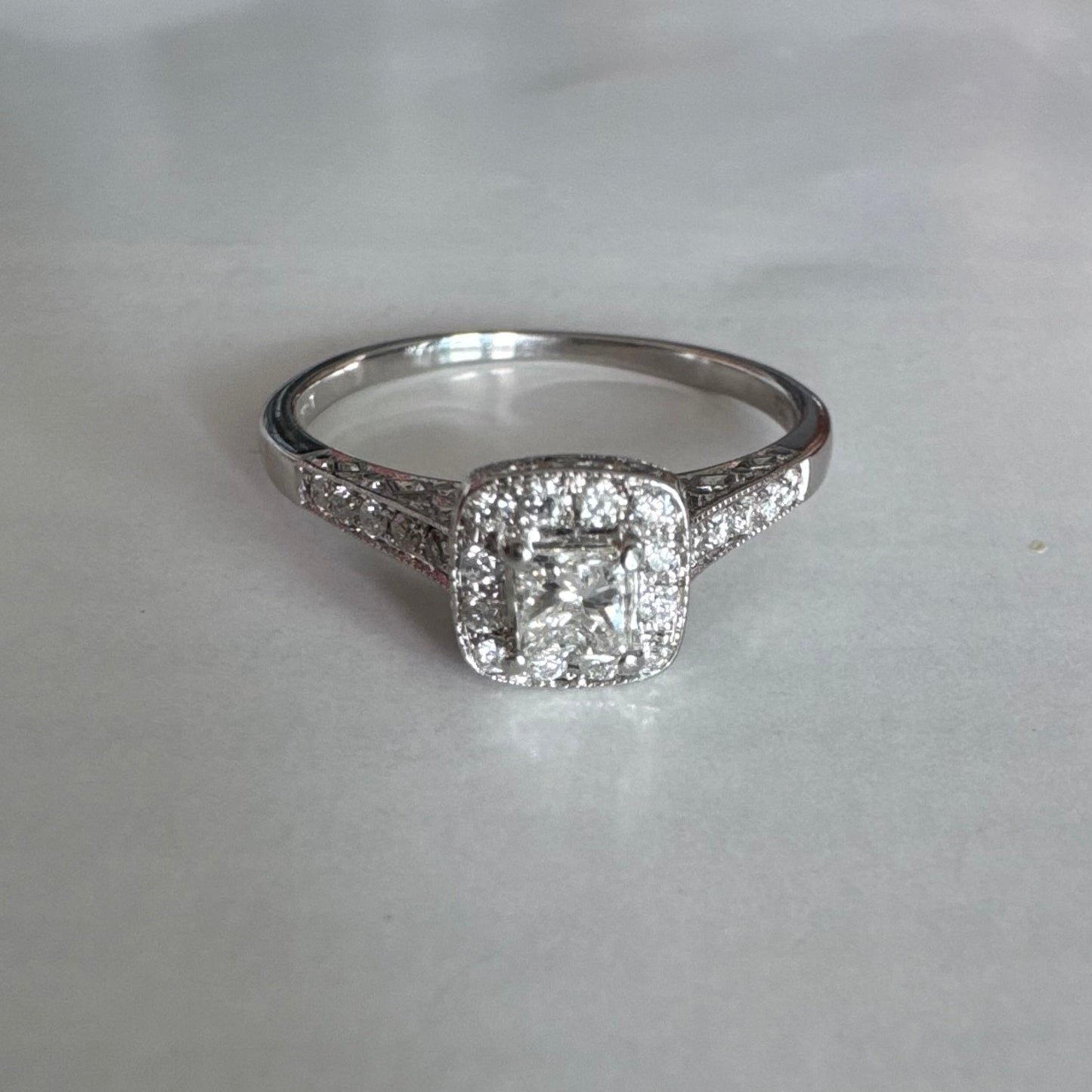 טבעת זהב לבן 14 קראט משובצת יהלום מרכזי בחיתוך פרינסס, מוקף הילת יהלומים קטנים וזרוע משובצת פאווה