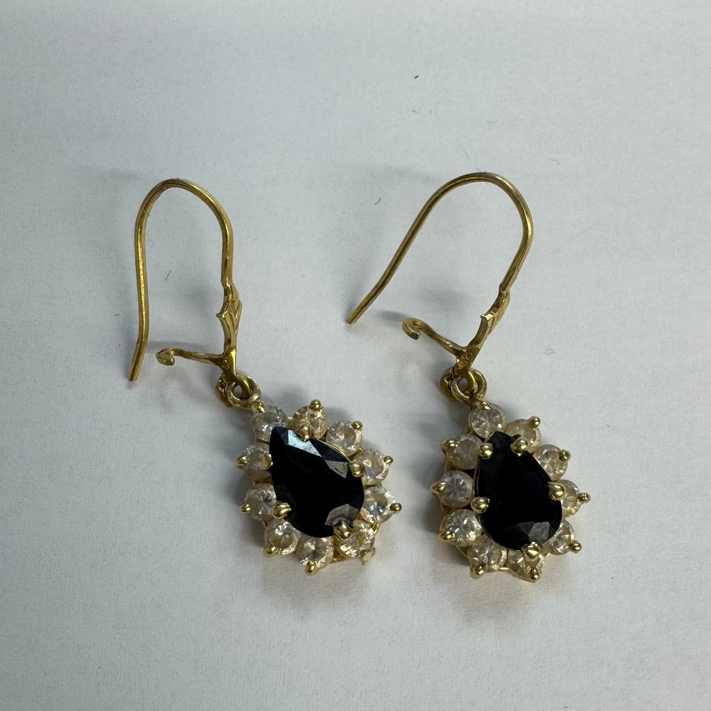 עגילי זהב צהוב 14 קראט בעיצוב קלאסי ומרשים, משובצים אבן ספיר בגוון כחול כהה בחיתוך טיפה ומסביבה אבנים שקופות עם הילה נוצצת שמוסיפה אור וזוהר לכל תנועה.